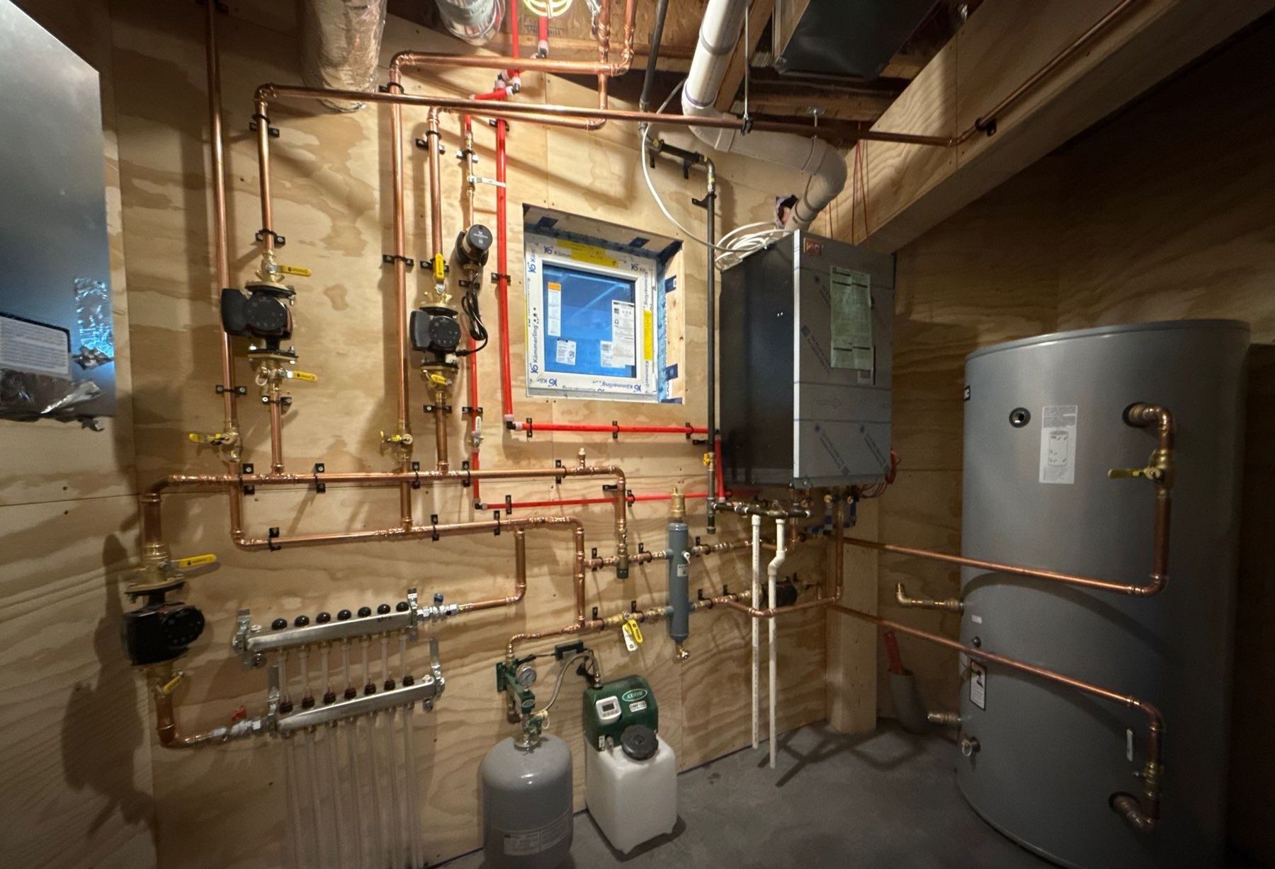 Finished mechanical room: copper headers and circulators on a plywood wall, radiant manifold, wall-hung boiler and a large indirect tank